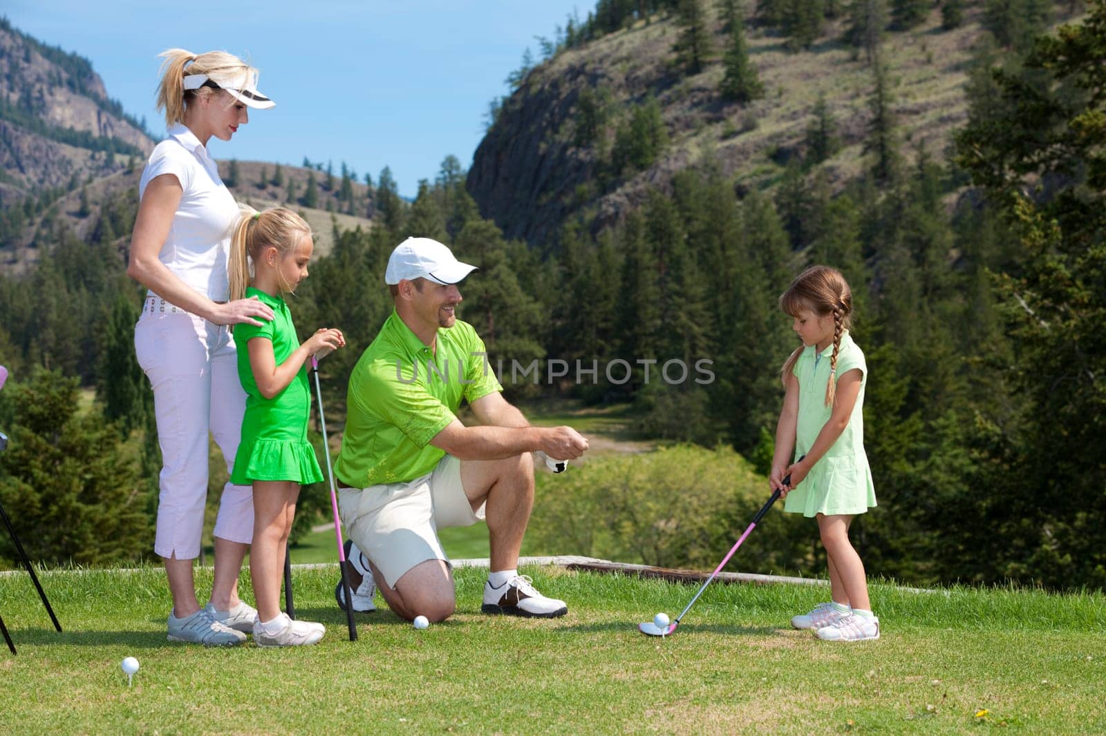 Outdoor photo of young family of four on golf course, father is instructing child.