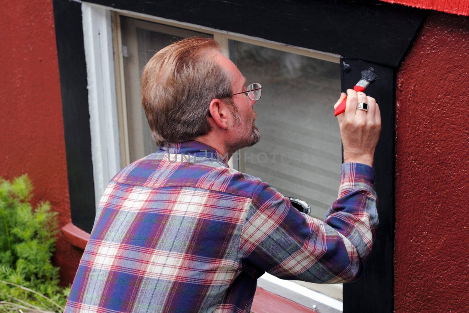 Man painting house window trim with black paint outdoors in the day.