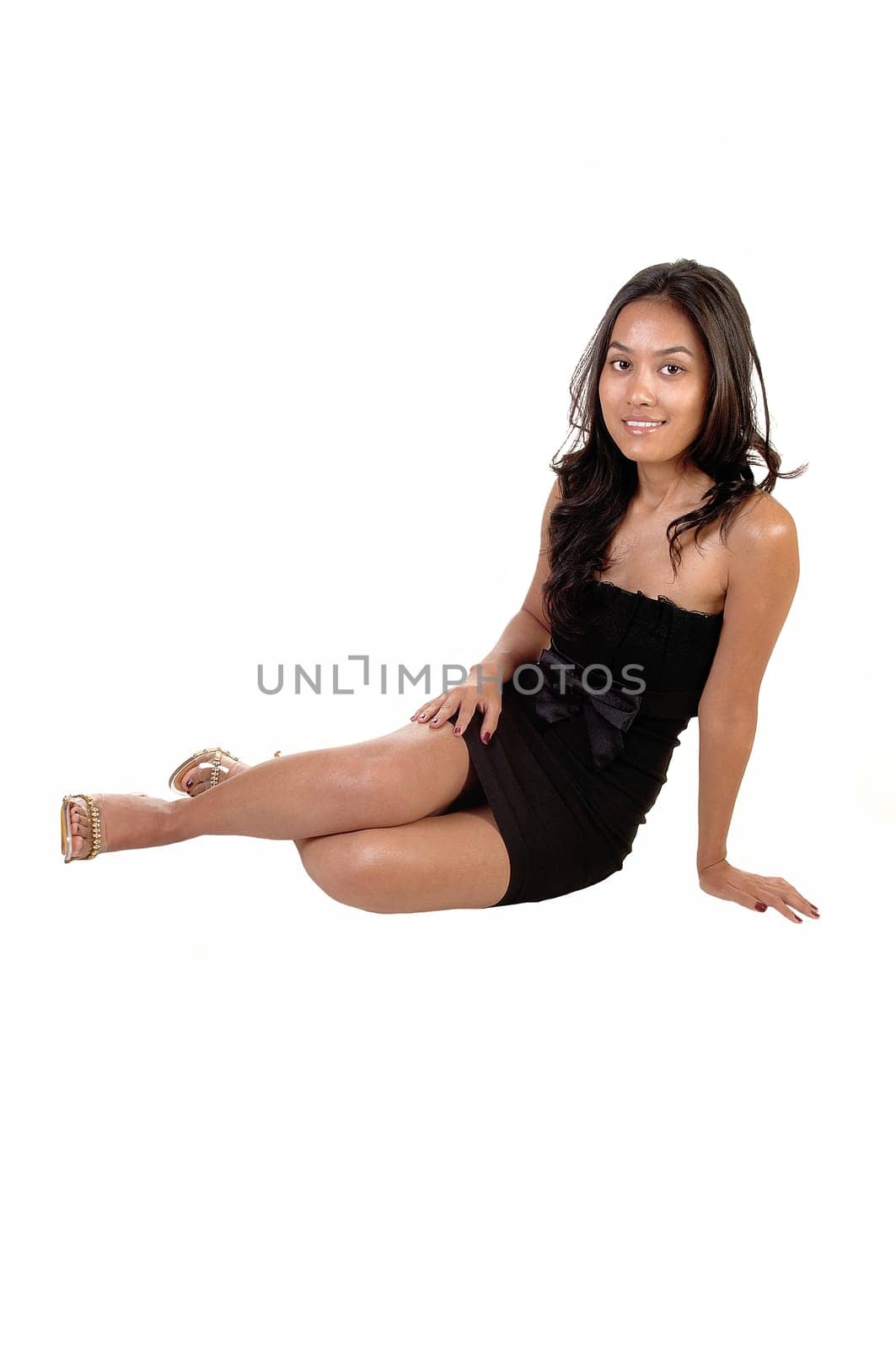 A pretty, slim Asian woman in a black dress with long curly brunette hair sitting on the floor and smiling into the camera, for white background.