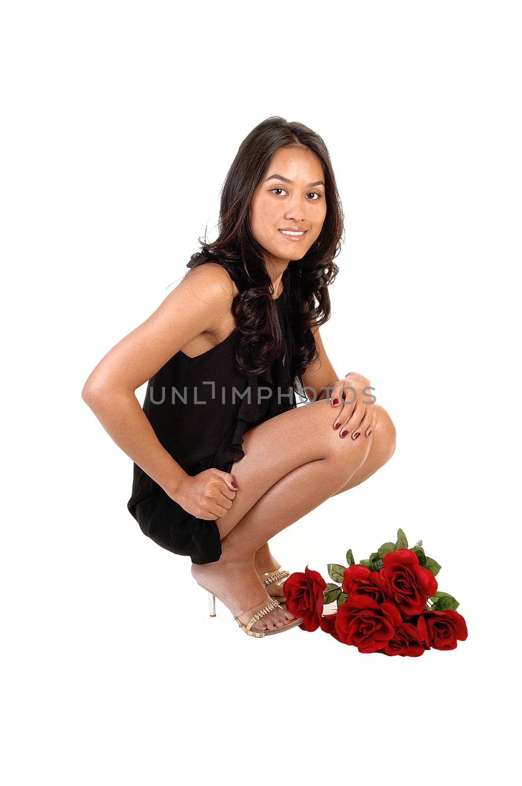 A lovely Asian girl crouching on the floor in a black dress and heels with a bunch of red roses to her feet, over white background.
