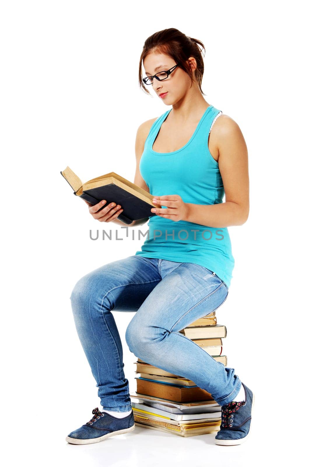 Young beautiful girl in glasses reading a book sitting on books. Isolated on white.