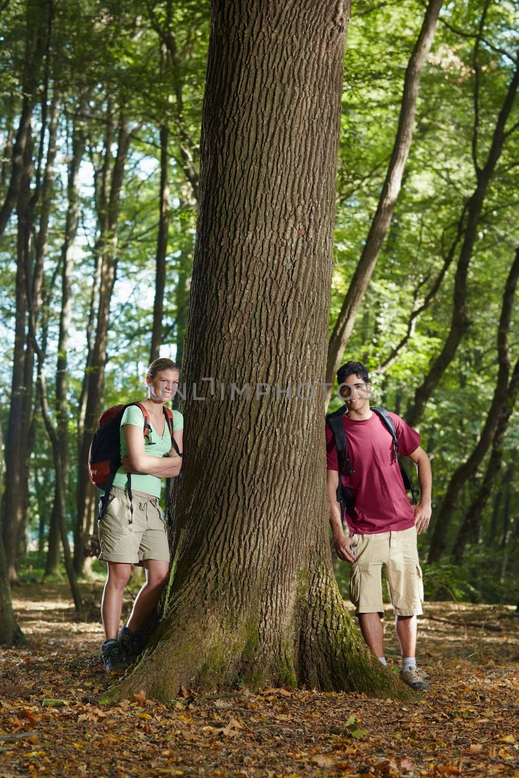 young man and woman during hiking excursion, posing near large tree and smiling. Vertical shape, front view, full length