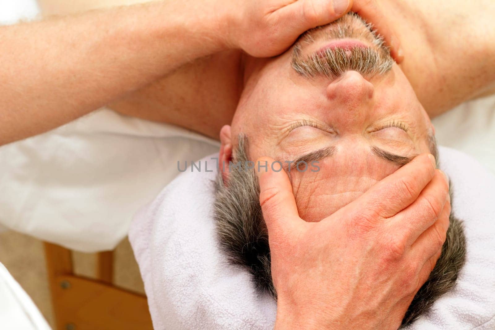 Mature Caucasian man in his 50s relaxing on a massage table with his eyes closed receiving a face massage from a massage therapist.