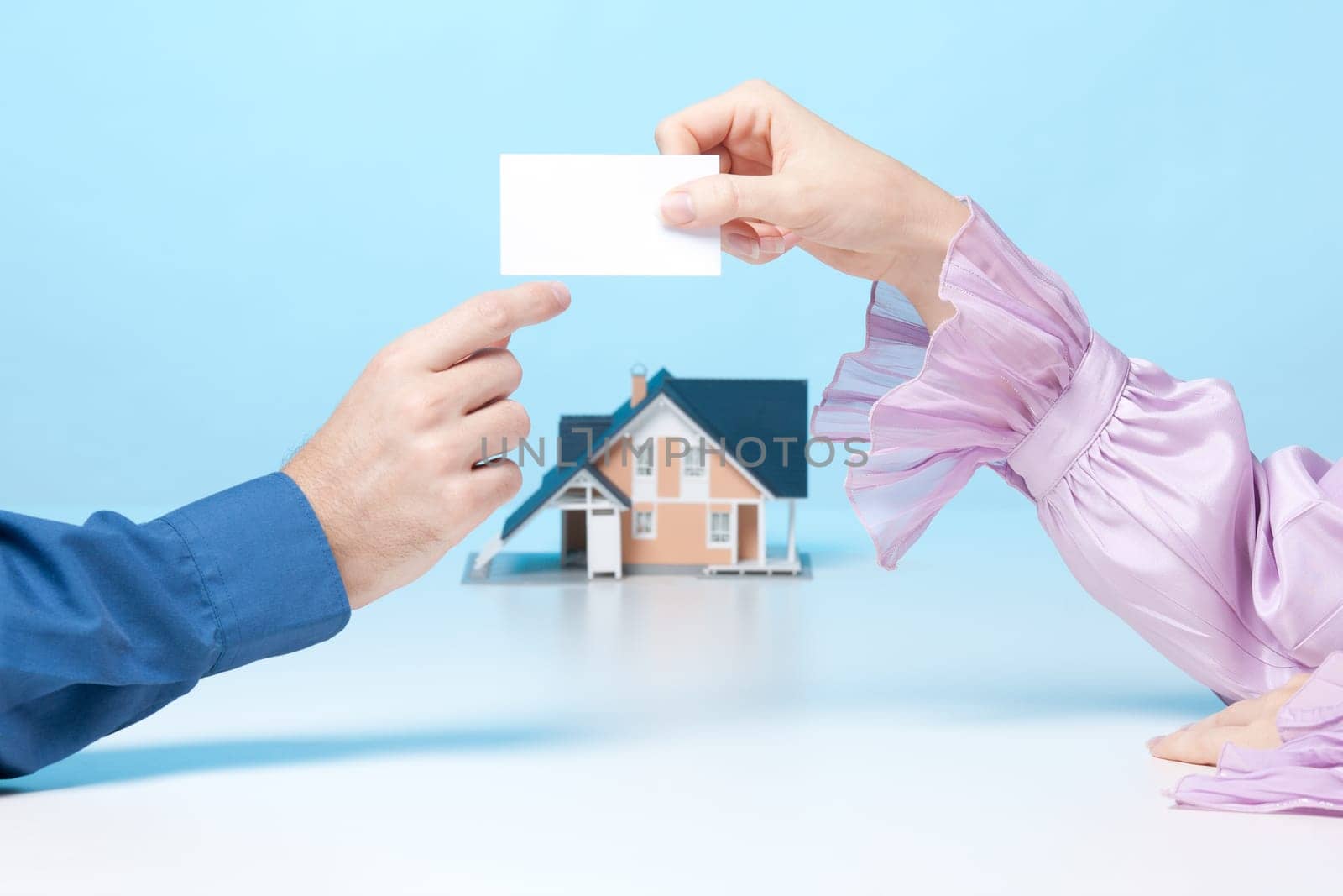 Real estate broker woman is giving to client her business card. Real estate agency is represented by model of house in background.