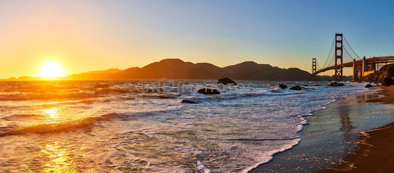 Image of Panorama sunset over waves against sandy shore and distant mountain with Golden Gate Bridge