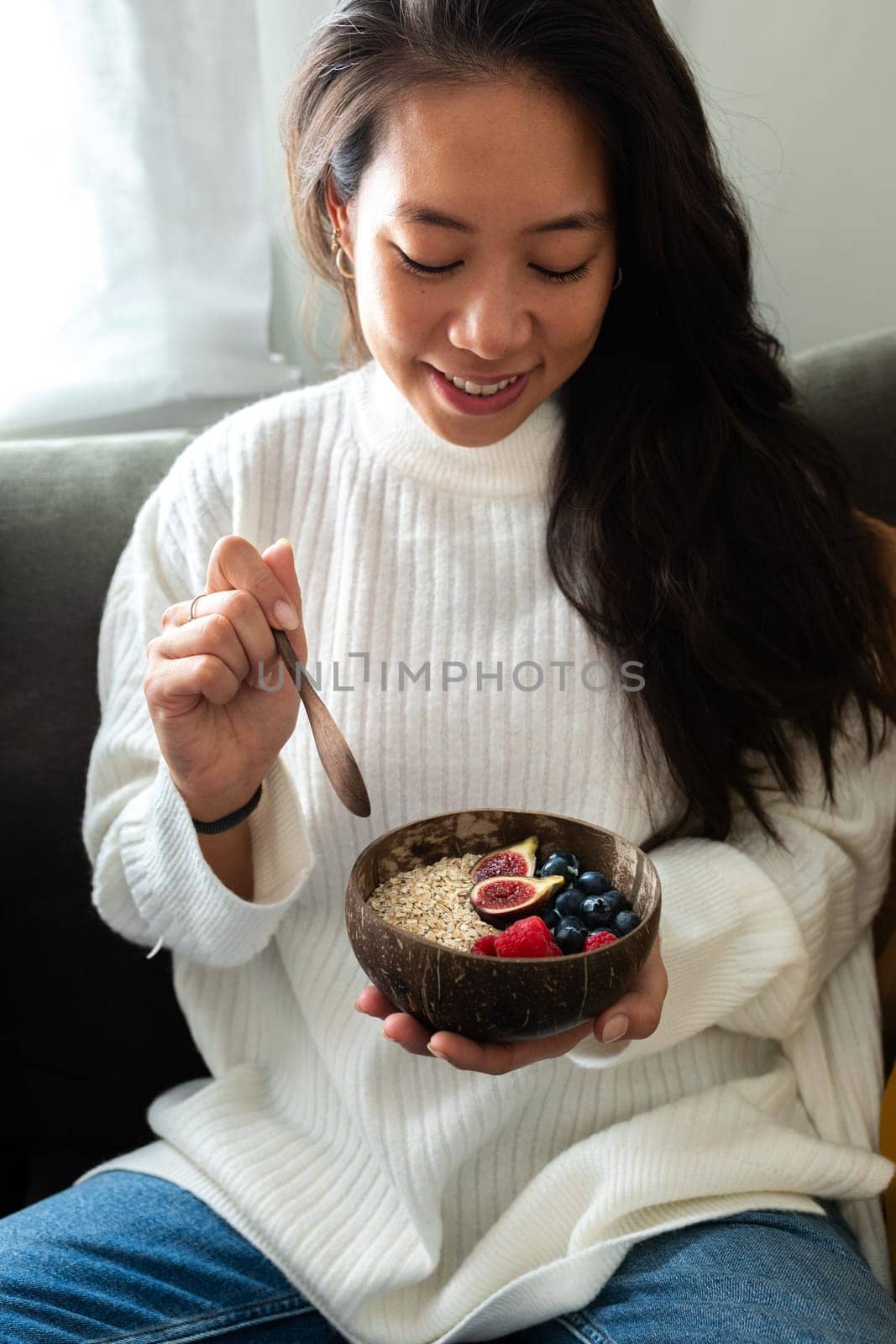 Vertical portrait of happy young Asian woman eating healthy smoothie breakfast bowl of oats and fruit relaxing on the couch. Healthy lifestyle concept.