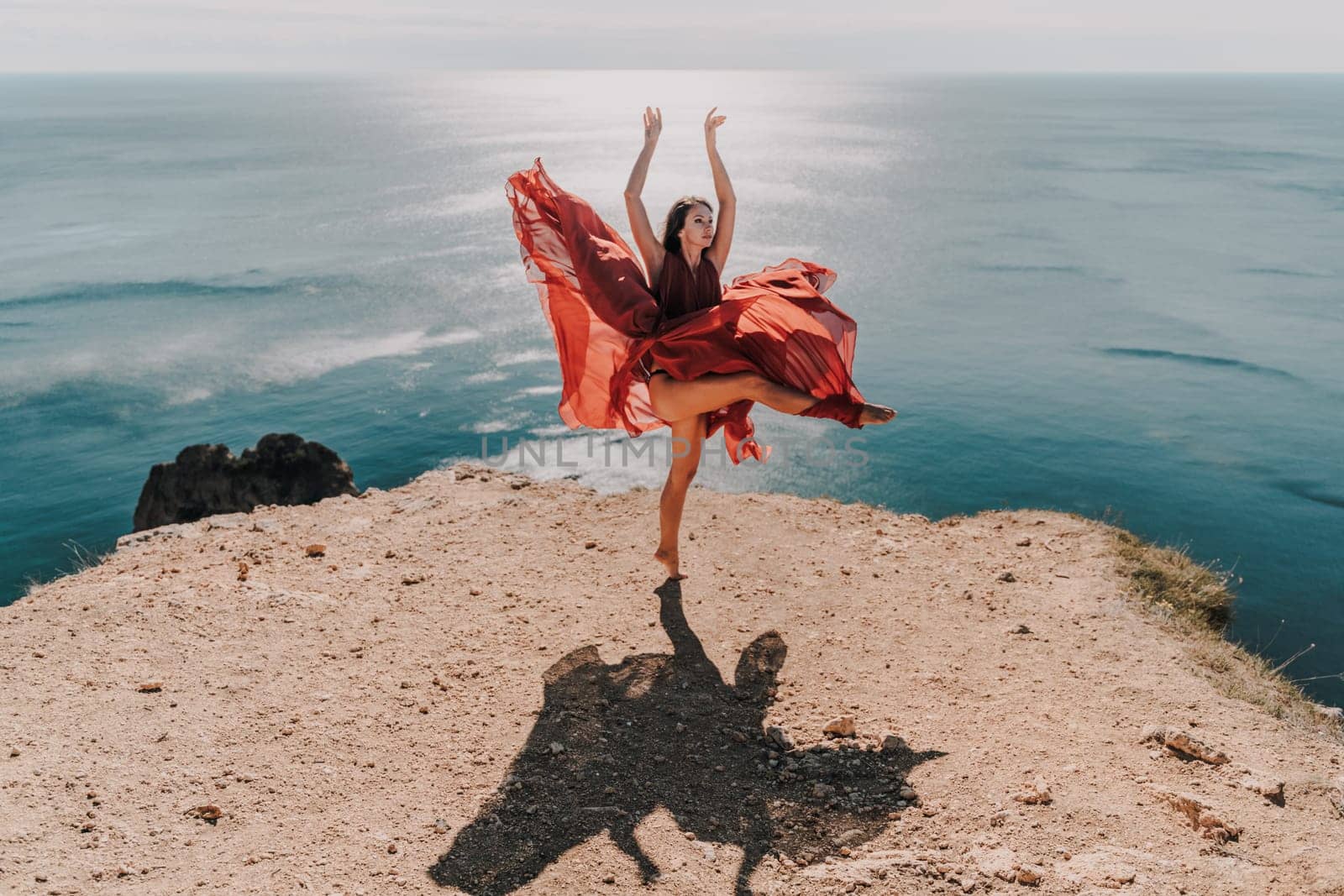 Woman red dress sea. Female dancer posing on a rocky outcrop high above the sea. Girl on the nature on blue sky background. Fashion photo. by Matiunina