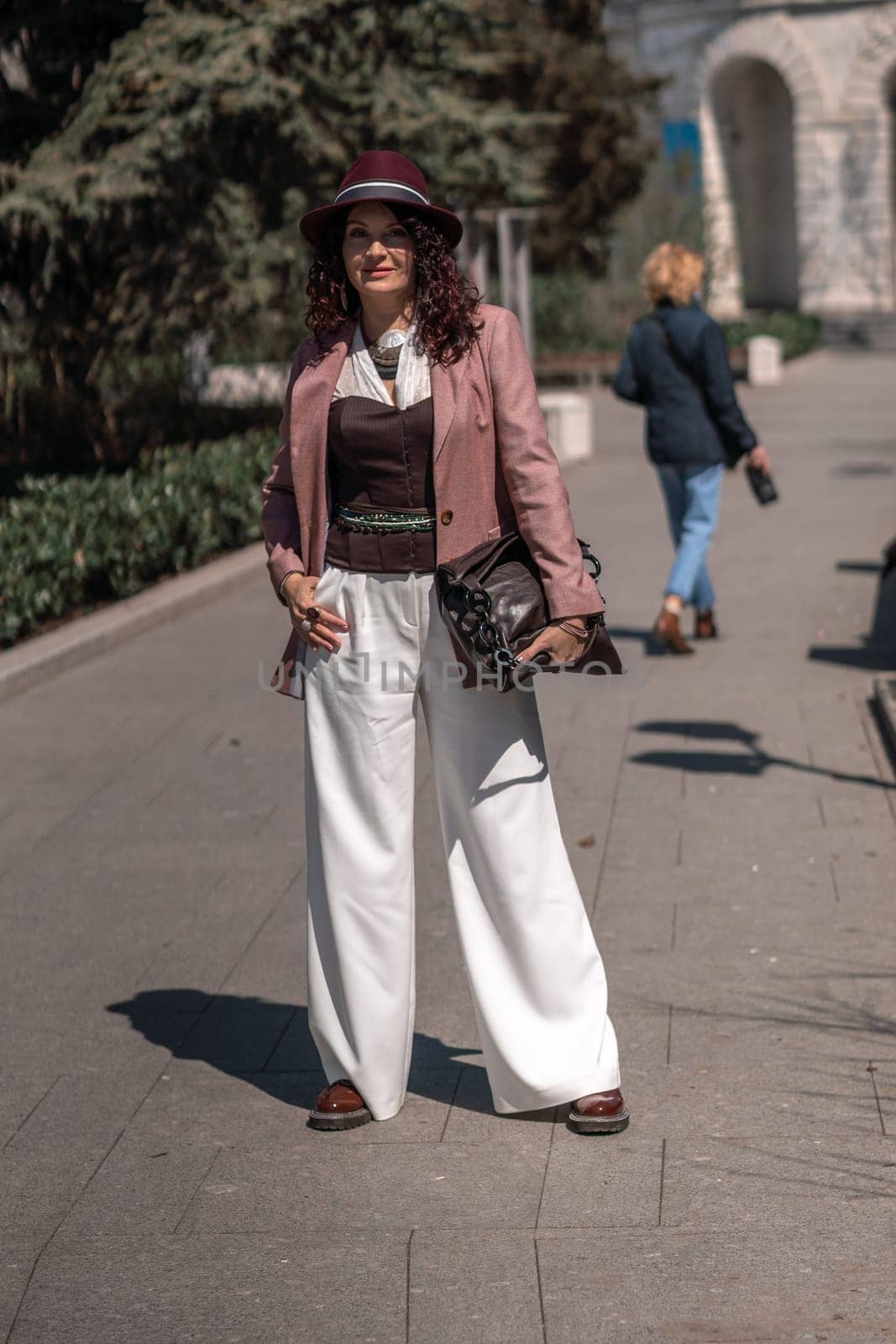 Woman park city. Stylish woman in a hat walks in a park in the city. Dressed in white corset trousers and a pink jacket with a bag in her hands. by Matiunina