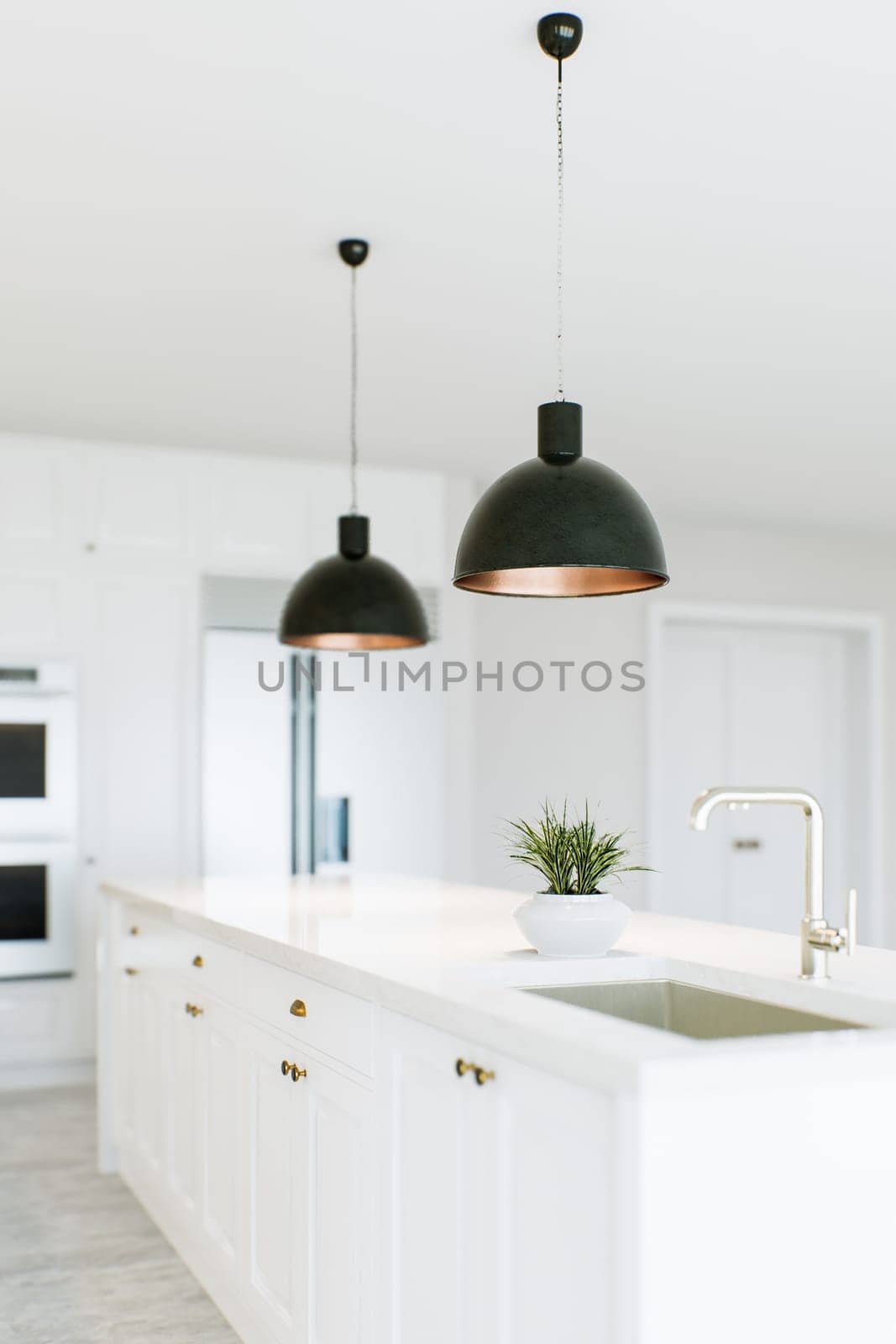 View of the kitchen island in a white kitchen. Dark lights over the island. Kitchen appliances in the background. Kitchen sink in island. 3D rendering.