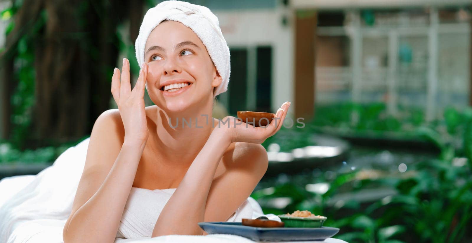 Beautiful caucasian women in white towel smells herbal homemade facial mask while lie on spa bed surrounded by outdoor natural environment. Relaxing and chilling concept. Tranquility.