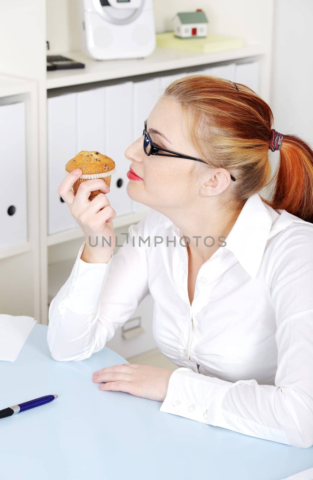 Beautiful caucasian woman in glasses smelling a cupcake in the office.