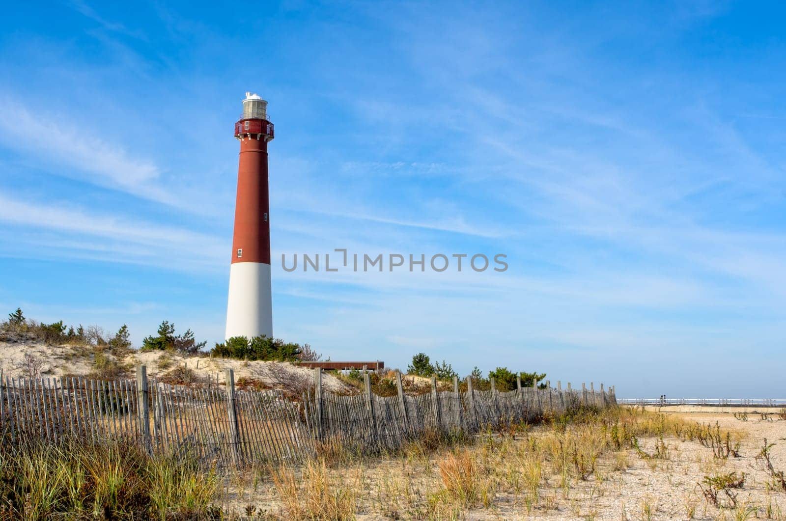 A lighthouse on the beach. There is also an old wooden fence on the beach in this landscape. This is Barnegat Lighthouse, nicknamed "Old Barney"", at Long Beach Island, New Jersey."