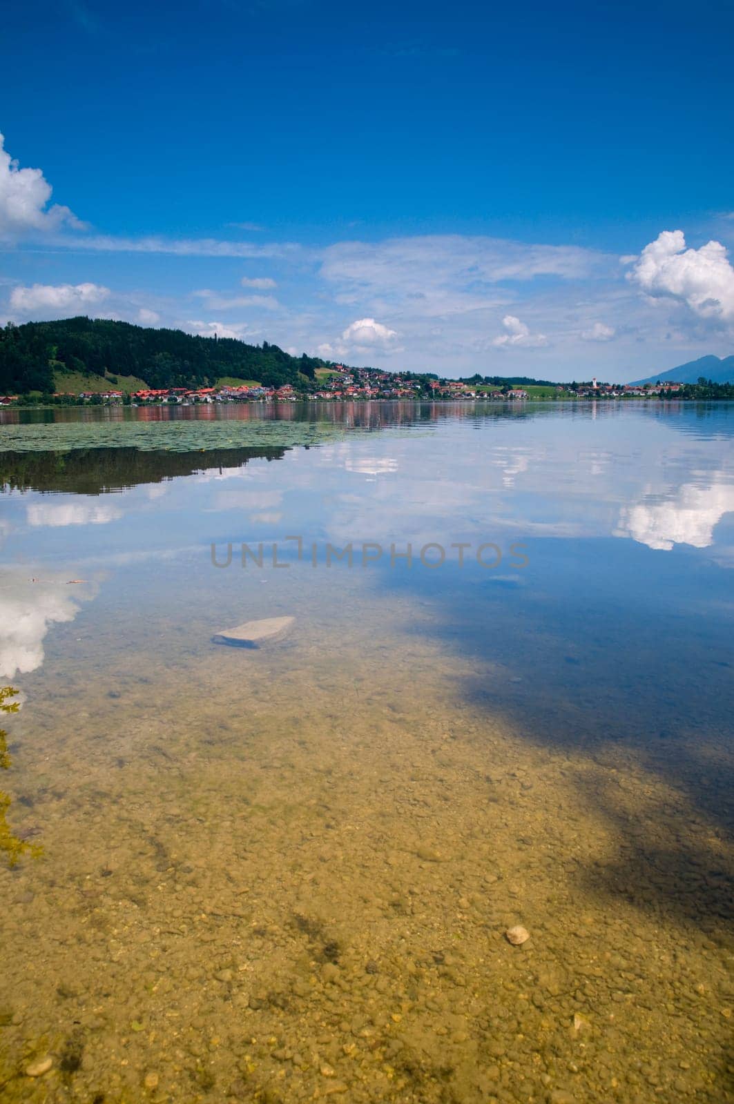 Wide-angle view of the Hopfensee with the community of Hopfen am See behind it under blue skies with small white clouds in the Allgäu / Bavaria in summer
