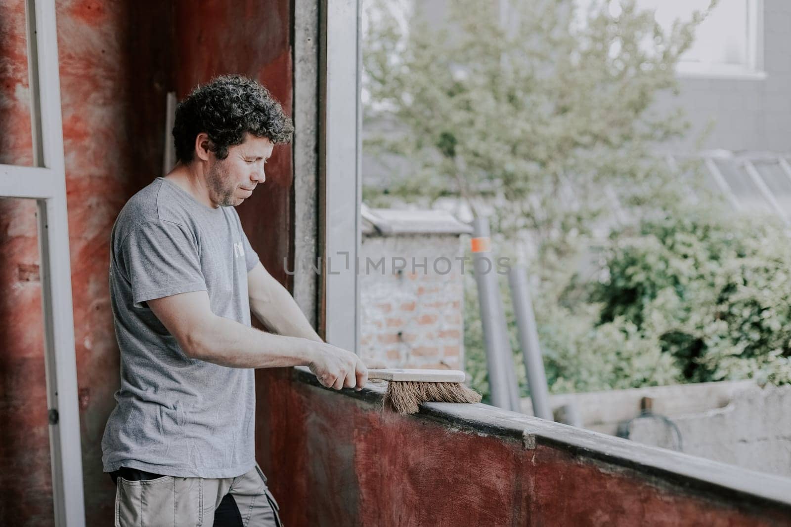 Young caucasian man holding an old broom and sweeping a dirty window frame while standing on the left, close-up side view. The concept of cleaning and installing windows, construction work, house renovation.