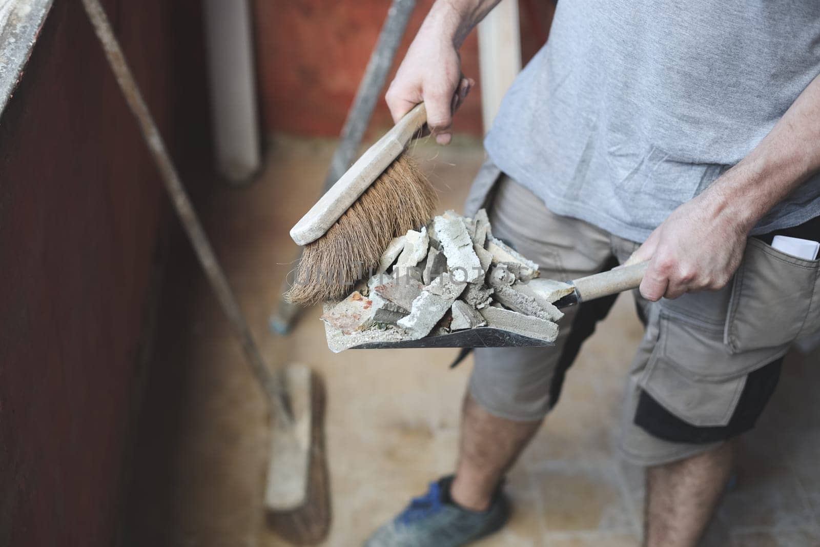 Young caucasian unrecognizable man holding a small broom of construction debris on an old shovel, close-up side view. The concept of cleaning and installing windows, construction work, home renovation.