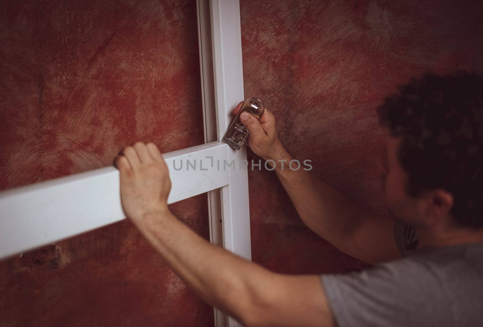 A young caucasian man with curly hair cleans the cracks on the window frame with a construction knife, preparing them for installation, close-up side view. The concept of home renovation, installation of windows, construction work.