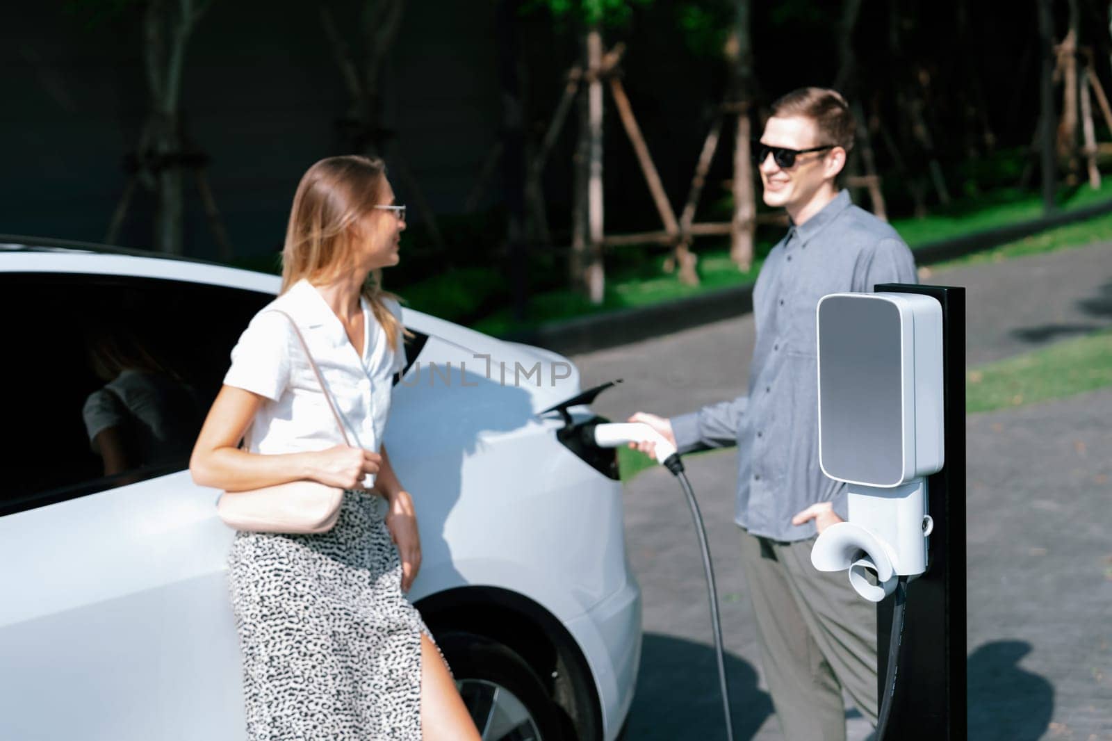 Young couple travel with EV electric car charging in green sustainable city outdoor garden in summer shows urban sustainability lifestyle by green clean rechargeable energy of electric vehicle innards