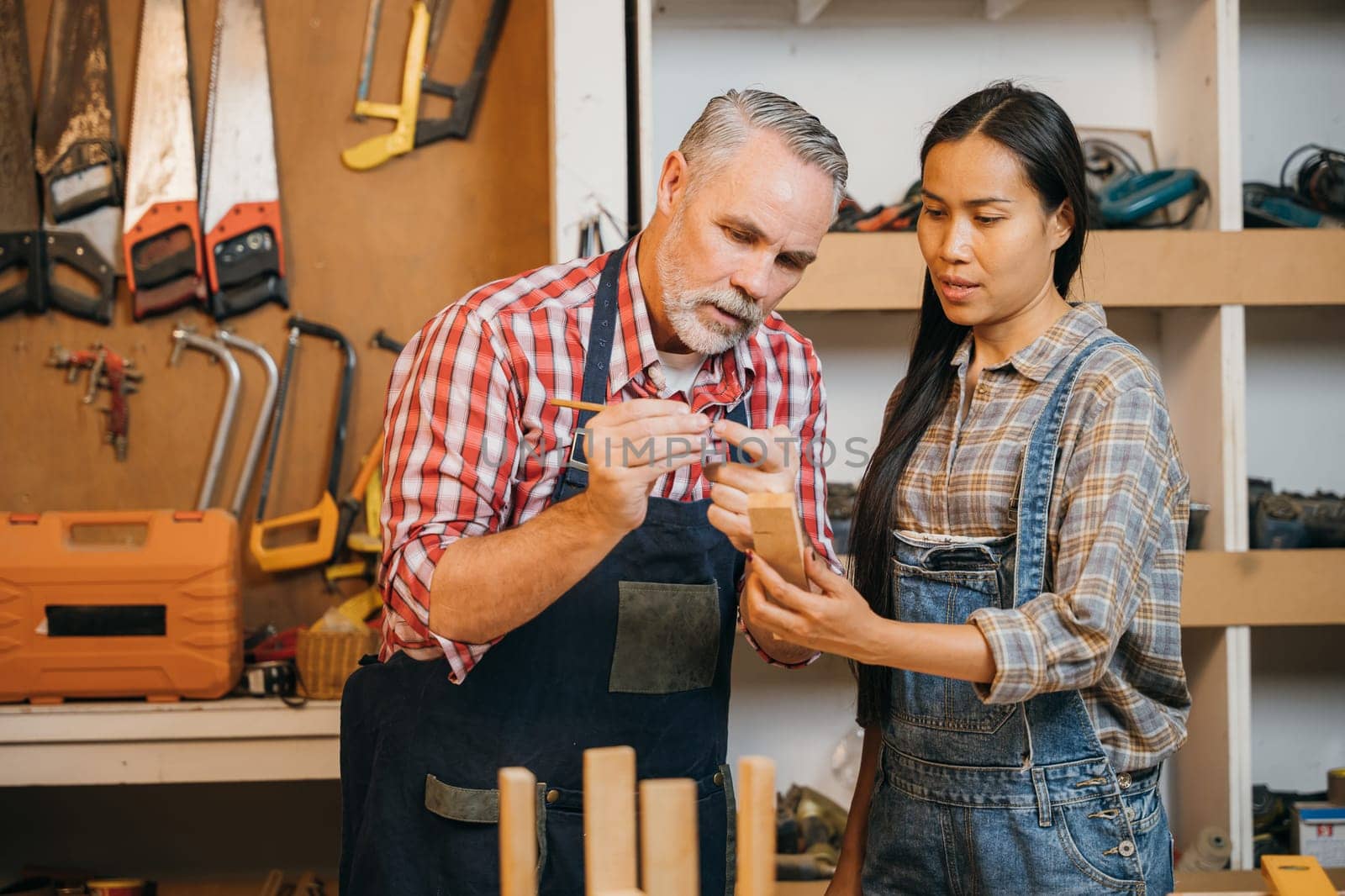 Craftsmen senior man teaching woman apprentice at woodshop, Carpenter training wood for female standing at wood table in workshop, National Carpenters Day