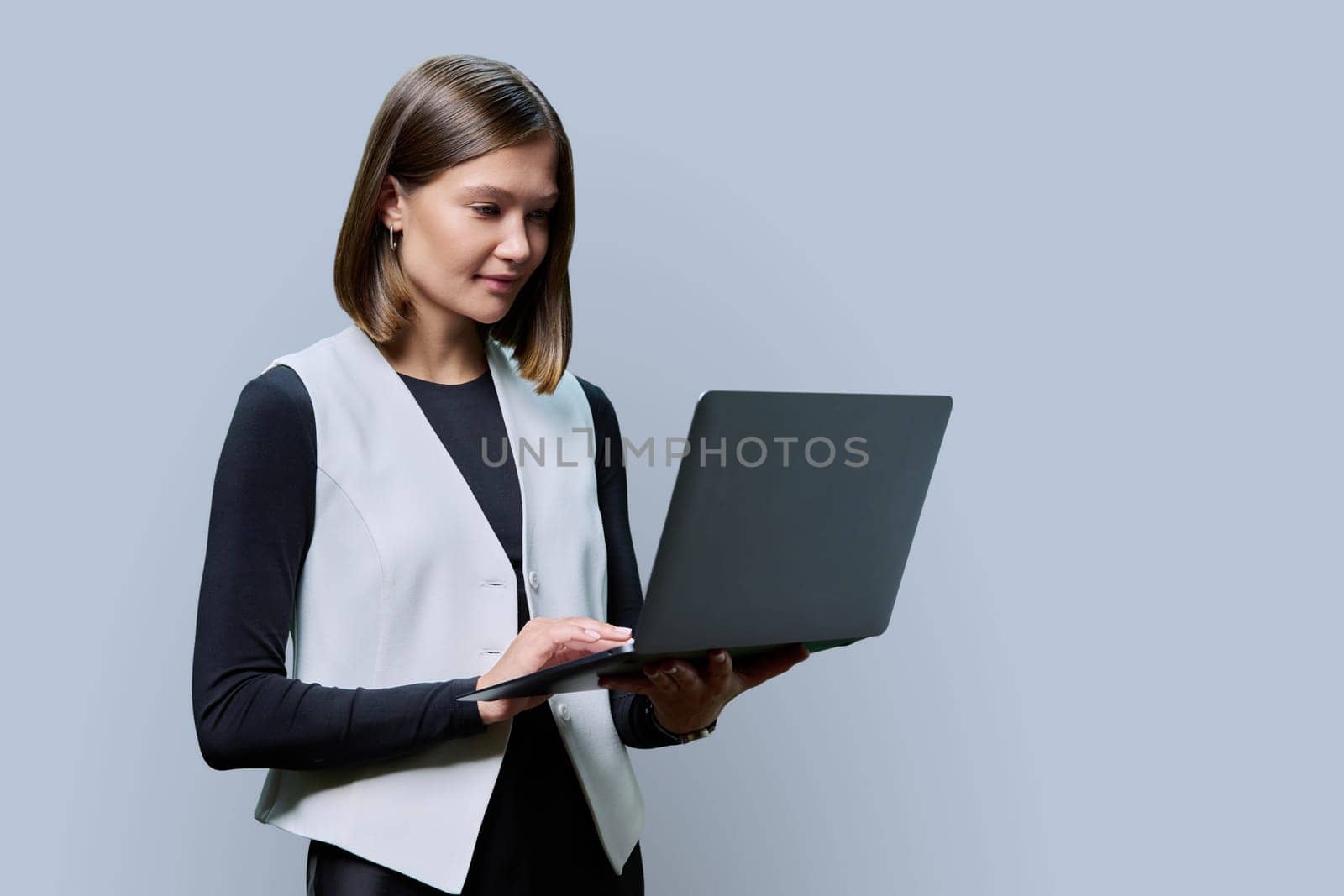 Young woman using computer on grey studio background. Serious female looking at laptop screen. Technologies in work business study leisure communication, online Internet applications, lifestyle people