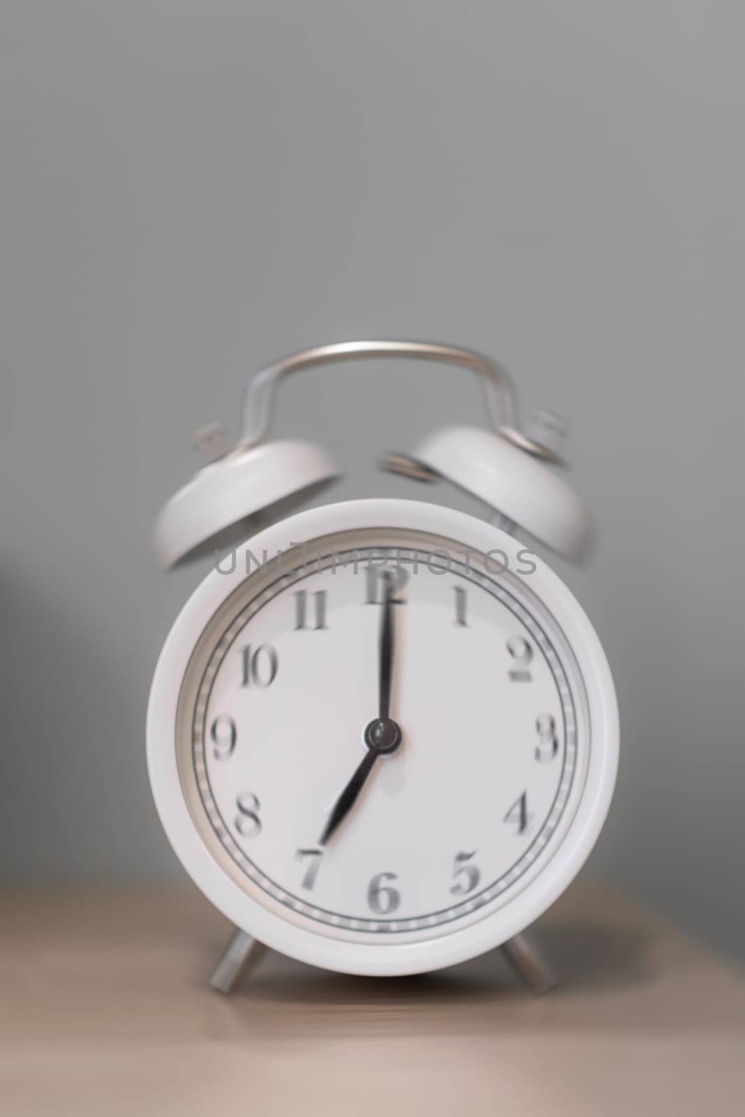 Close-up of a round white alarm clock on a table in the bedroom. Blurred photo. The hands on the clock show seven o'clock in the morning, time to get up. Retro alarm clock on the table, vintage tone.
