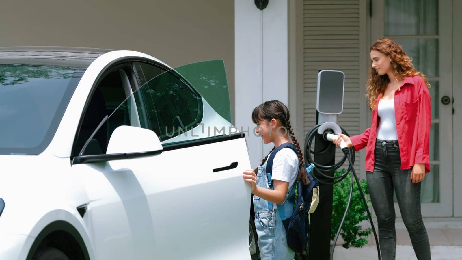 Little young girl help her mother recharge EV car at home. Synchronos by biancoblue