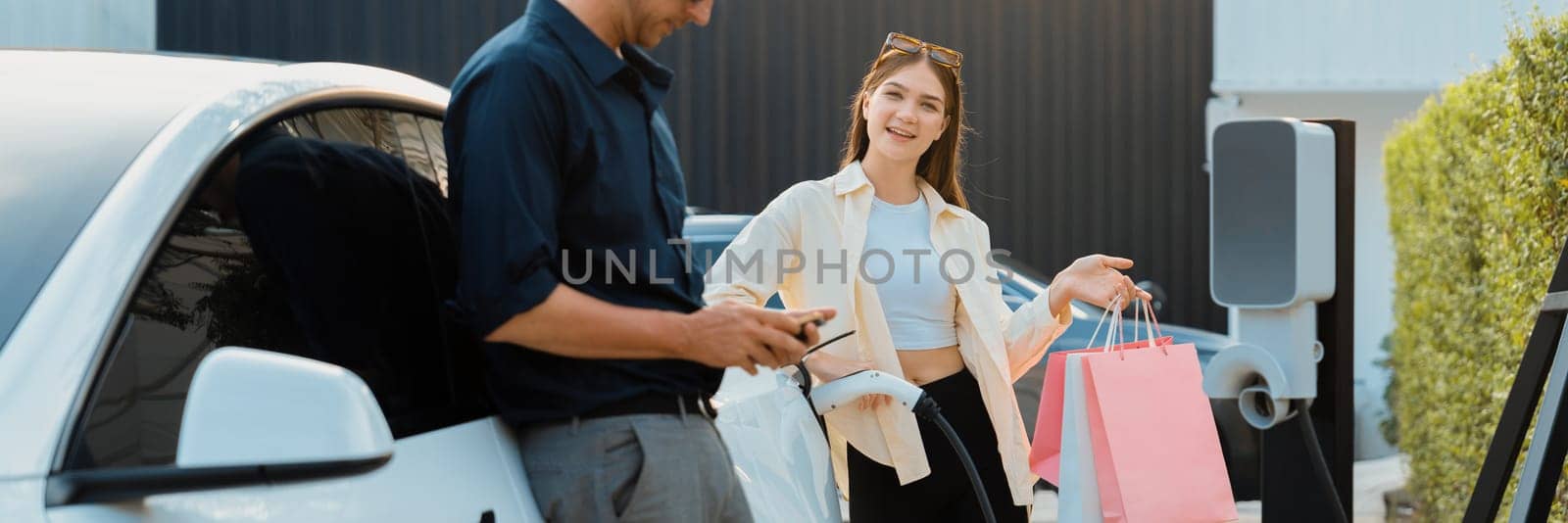 Young couple with shopping bag use smartphone to pay for electricity to recharge EV car battery from charging station at city mall parking lot. Happy couple go shopping by eco car.Panorama Expedient