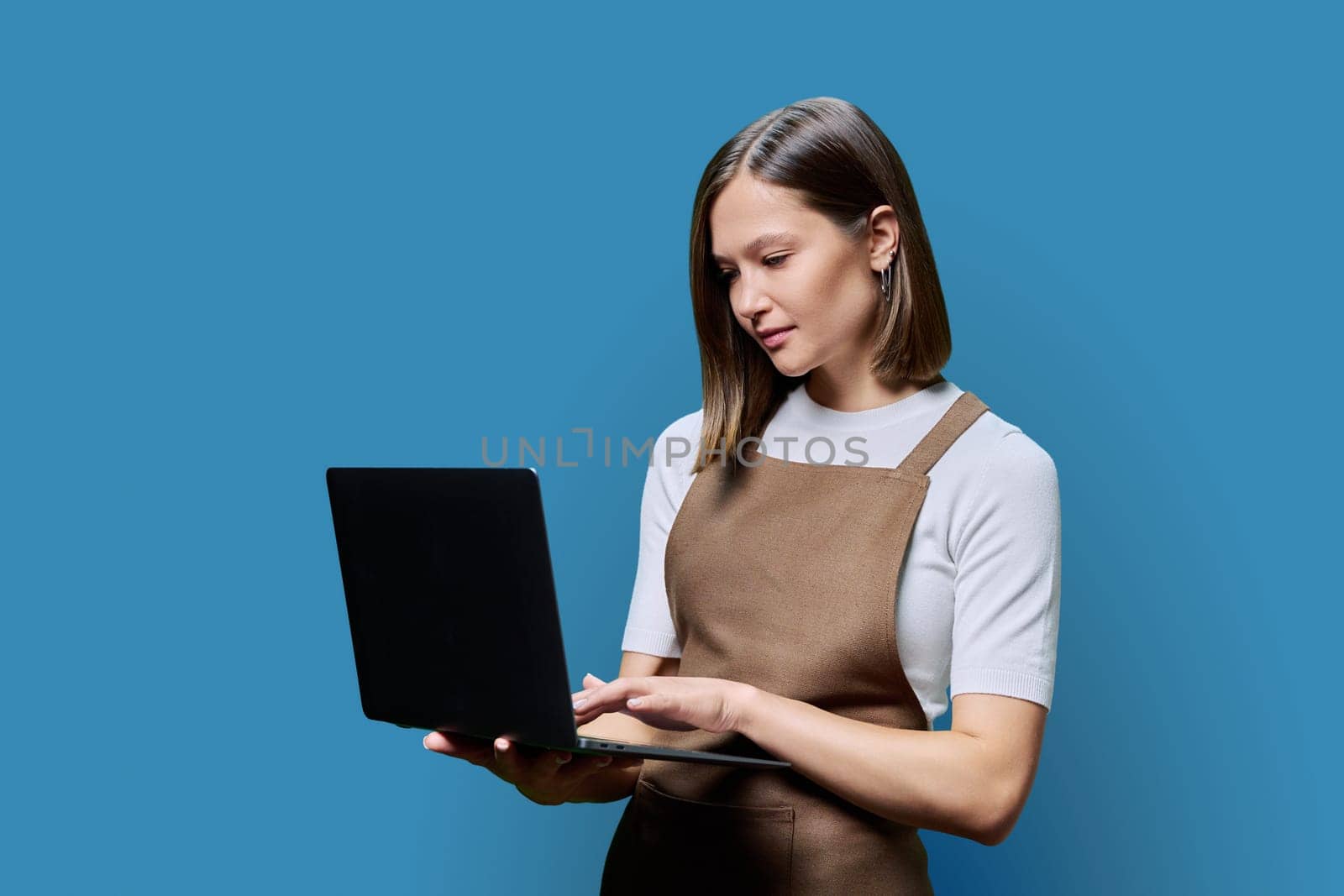 Portrait of young serious confident woman in an apron using laptop, looking at computer screen on blue studio background. Worker, startup, small business, job, service sector, staff, youth concept