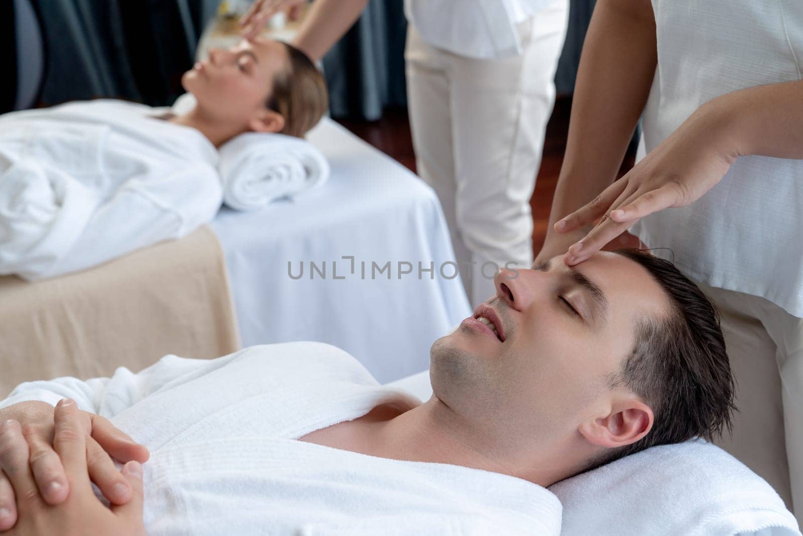Caucasian couple enjoying relaxing anti-stress head massage and pampering facial beauty skin recreation leisure in dayspa modern light ambient at luxury resort or hotel spa salon. Quiescent