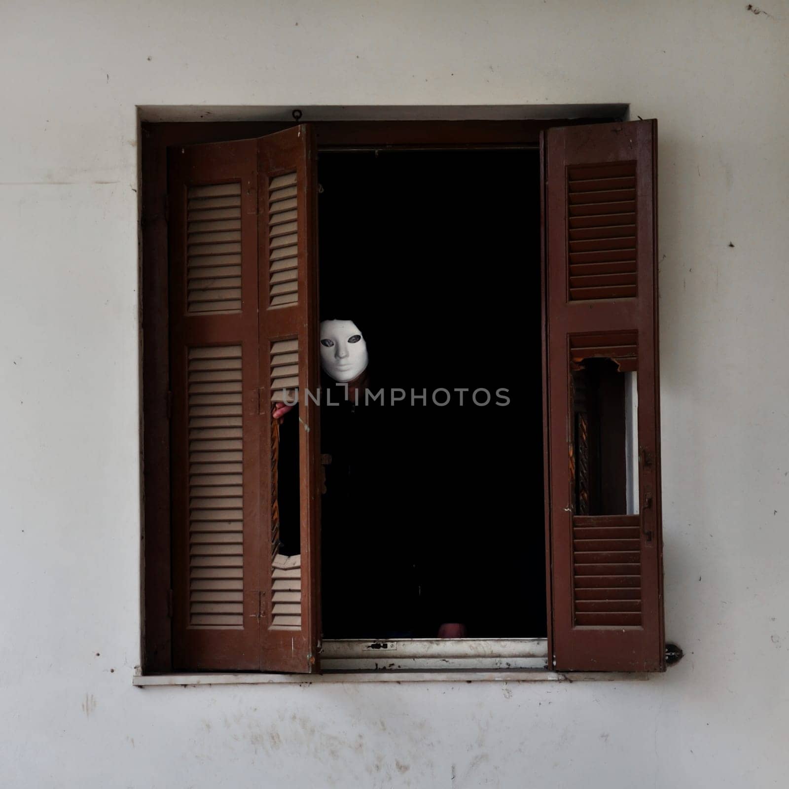 Man with white mask by the broken window shutter of an abandoned house.