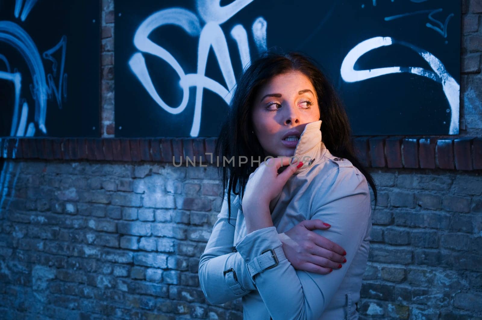 young black-haired woman in trench coat alone in the evening on a street with graffiti on a brick wall