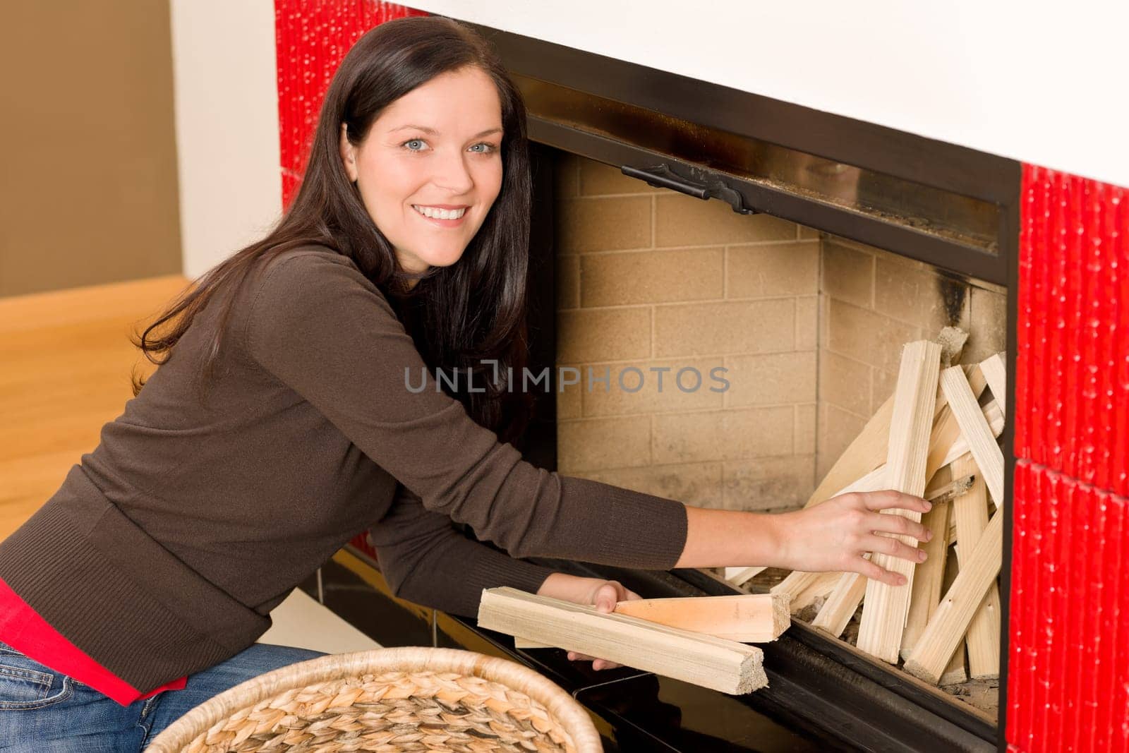 Young woman putting wood logs from wickerwork basket into fireplace