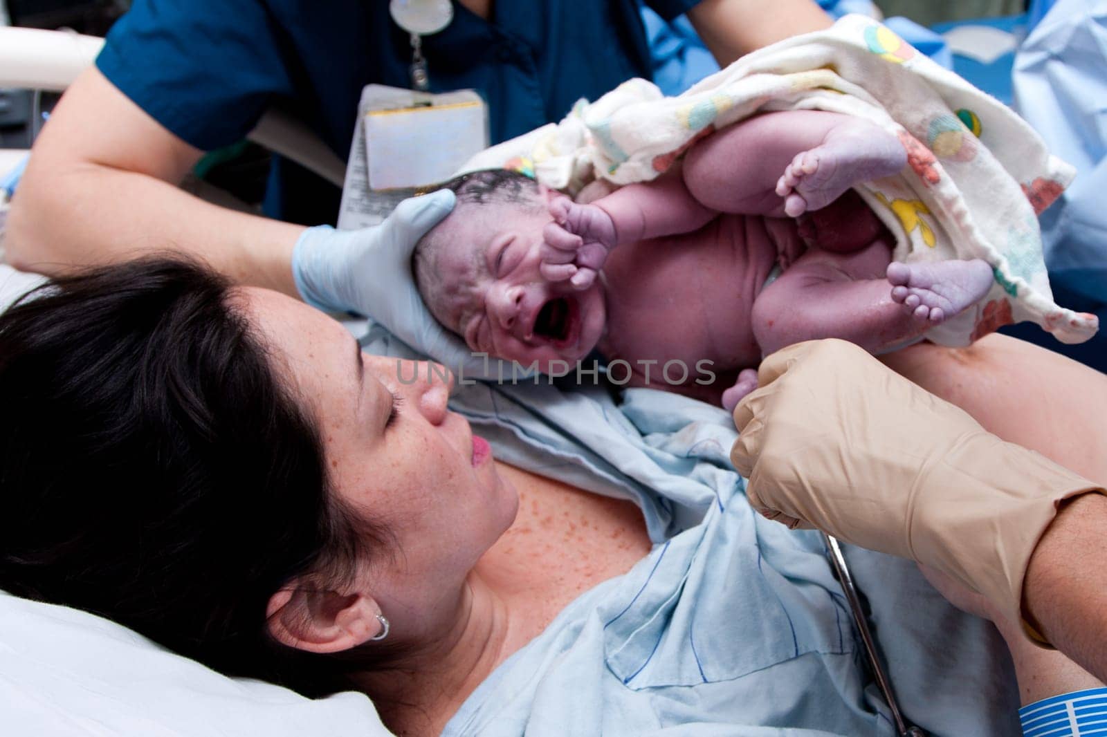 Mother in labor giving birth to baby new life in hospital. Infant covered in grease and lanugo is crying and held by nurses and doctors.
