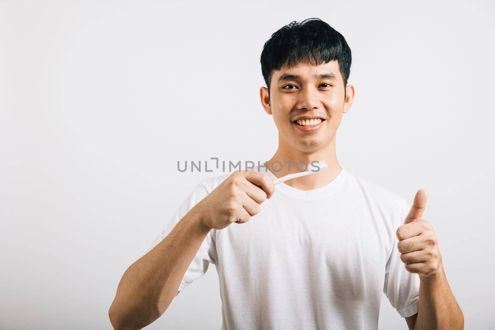 A confident Asian teenager promotes dental health by brushing his teeth with a smile and a thumbs-up. Studio shot isolated on white, emphasizing dental care and oral hygiene.