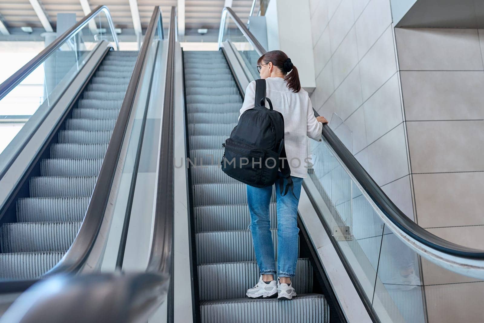 Middle-aged woman with backpack in casual style jeans on an escalator, in modern station building, airport, back view