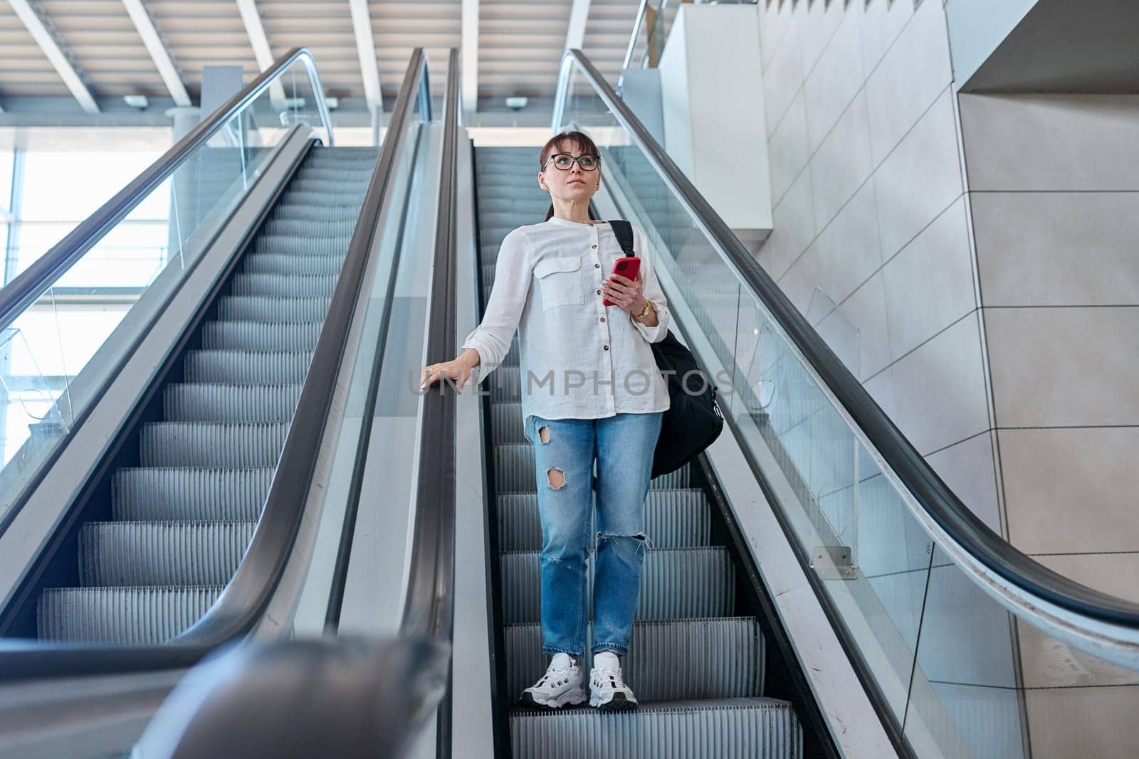 Middle-aged woman with backpack in casual style jeans on an escalator, in modern station building, airport
