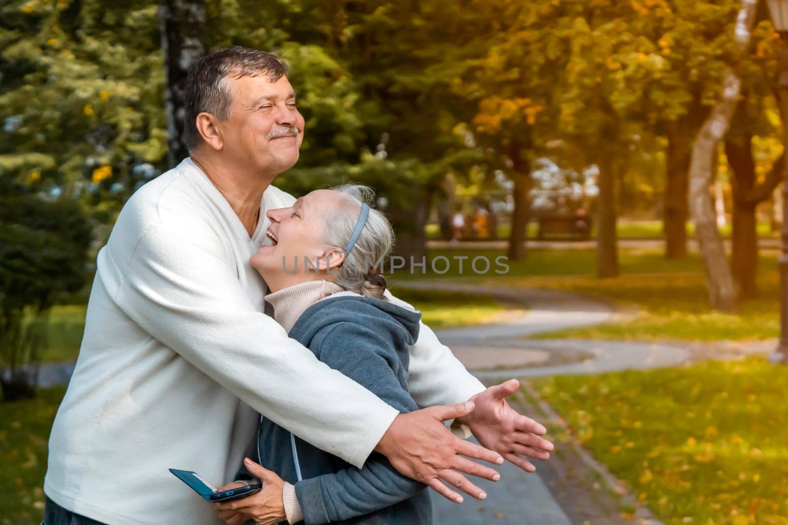 Happy elderly couple have fun together on a walk in the autumn park, loving caring husband laughs and hugs his wife, funny man and woman enjoy their leisure time at an older age.
