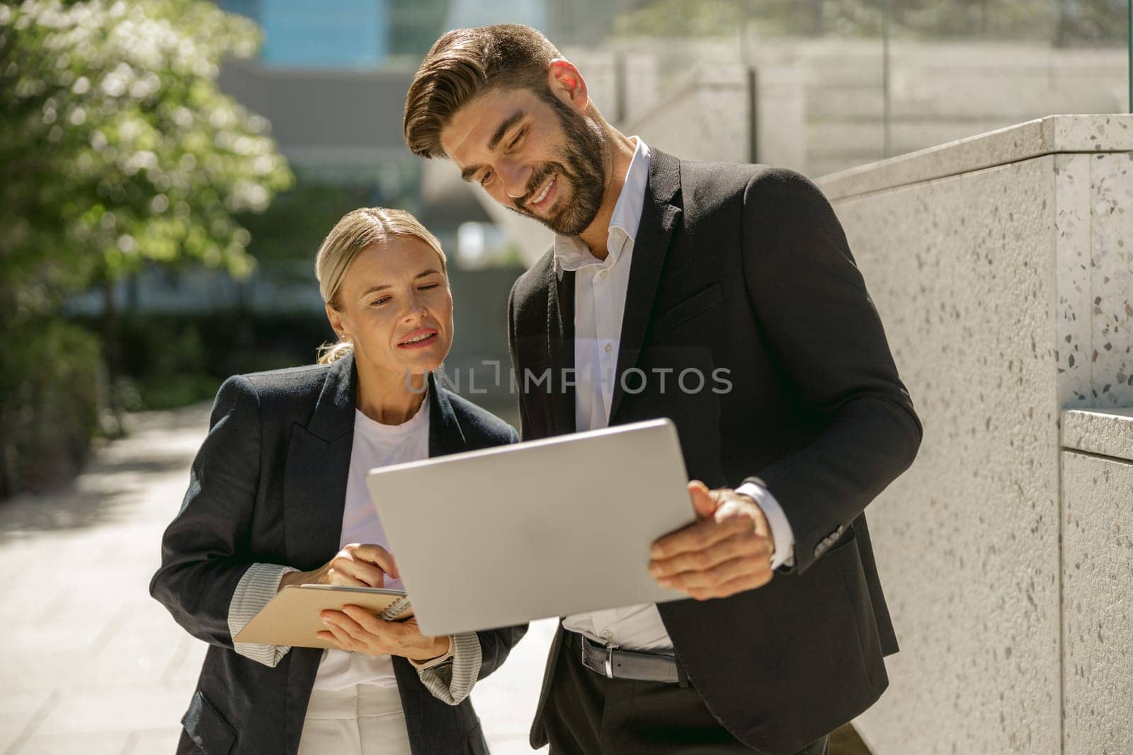 Man and woman in classic suit discussing business details and using laptop while standing outdoors