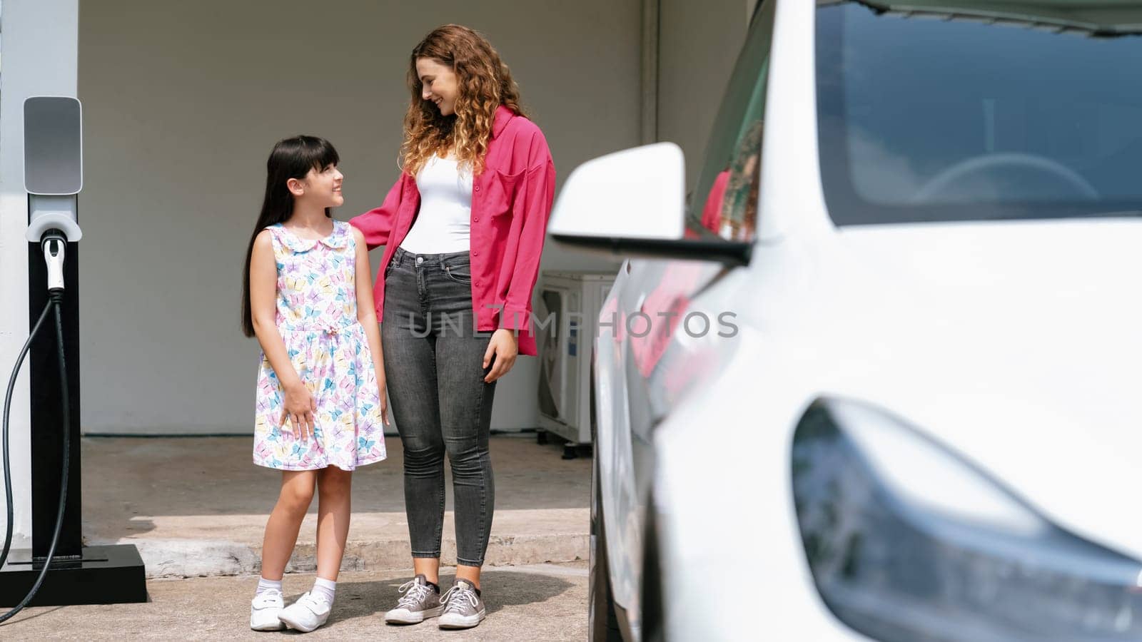 Happy little young girl learn about eco-friendly and energy sustainability as she help her mother recharge electric vehicle from home EV charging station. EV car and modern family. Panorama Synchronos