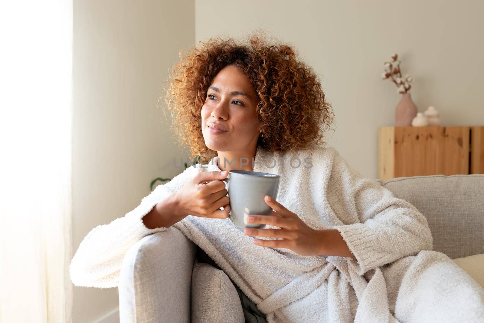 Pensive multiracial woman relaxing at home, sitting on the sofa drinking tea. by Hoverstock