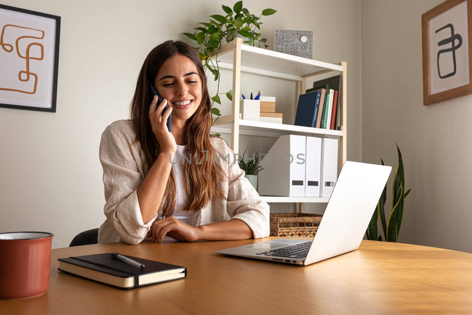 Happy young woman talking on the phone while working at home office using laptop. Technology concept.