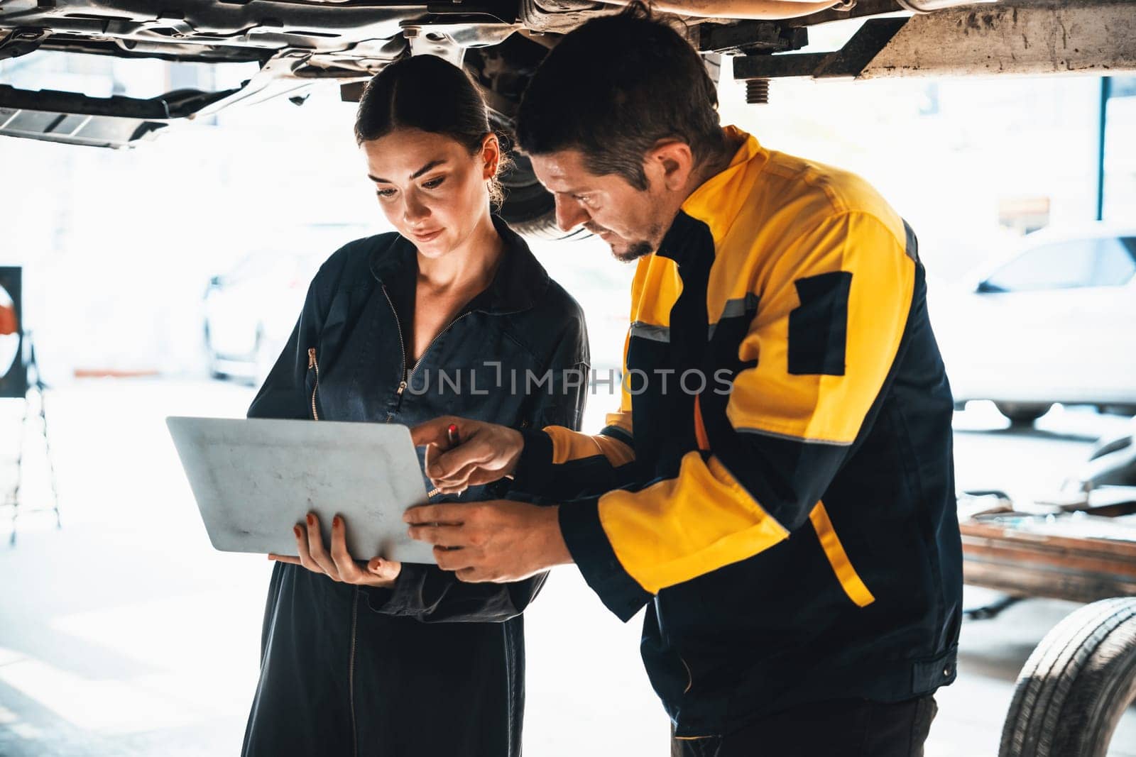 Two vehicle mechanic working together underneath lifted car, conduct car inspection with laptop. Automotive service technician in uniform carefully make diagnostic troubleshooting. Oxus