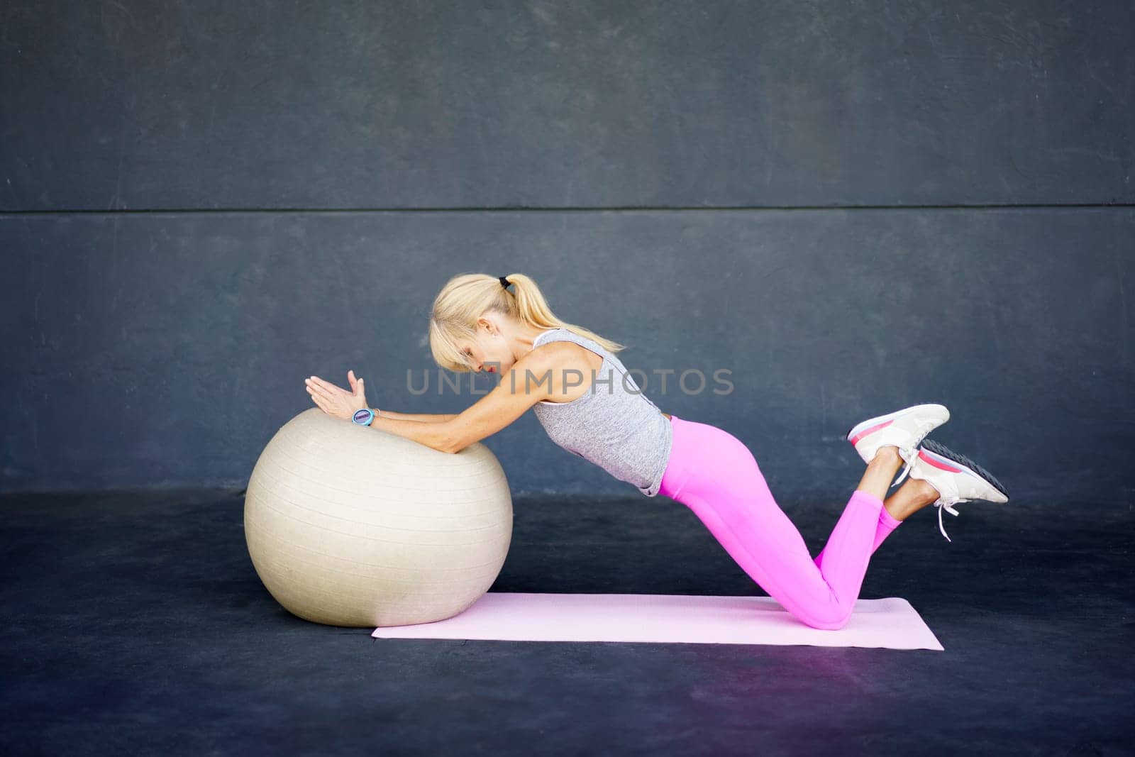 Side view of adult female in sportswear, ponytail looking down while lying with stretched hands on fit ball on gym floor and knees of folded up legs on mat in daylight