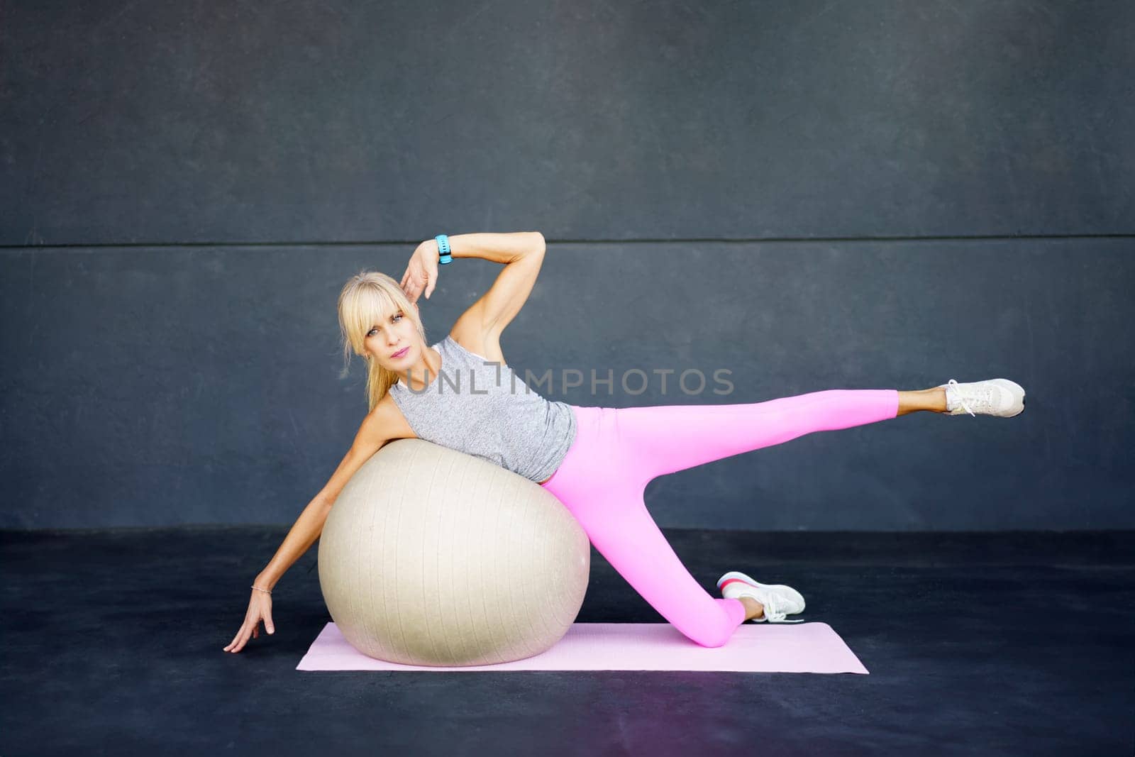 Adult female in sportswear sneakers looking at camera, while lying on side on fitness ball on mat with leg raised fingers touching gym floor and hand near head in daylight