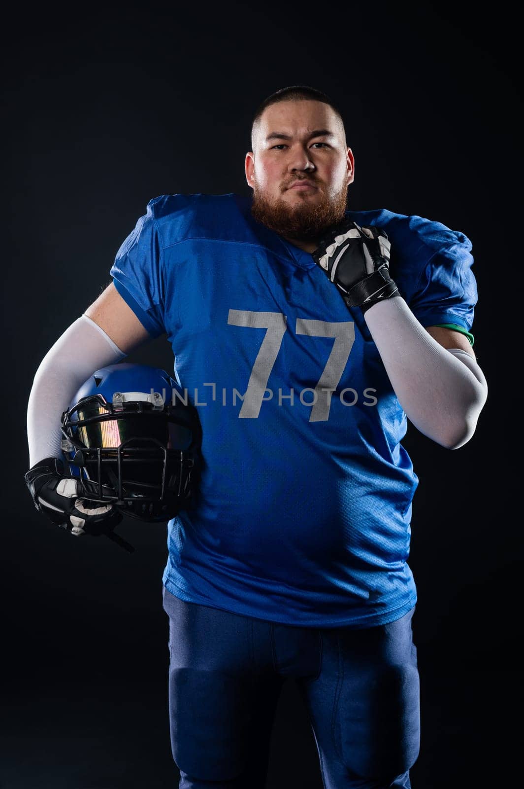 An Asian man with a red beard in a blue american football uniform holds a helmet on a black background. by mrwed54