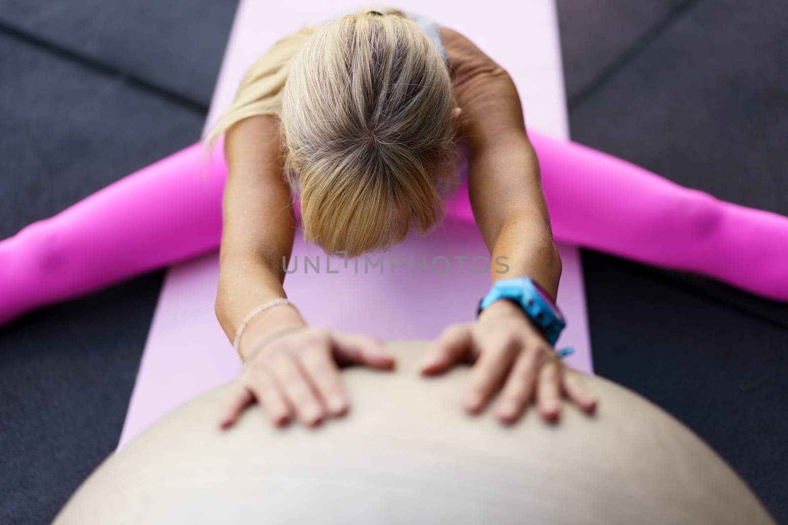 High angle of anonymous female athlete with smartwatch, looking down while sitting with legs apart on mat with hands on fit ball and doing stretching exercise in gym in daylight