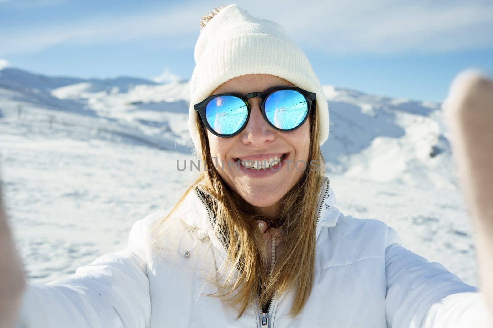 Positive young female in warm clothes beanie cap, and polarized sunglasses smiling and looking at camera while taking selfie and standing on snowy mountain slope on sunny day in winter