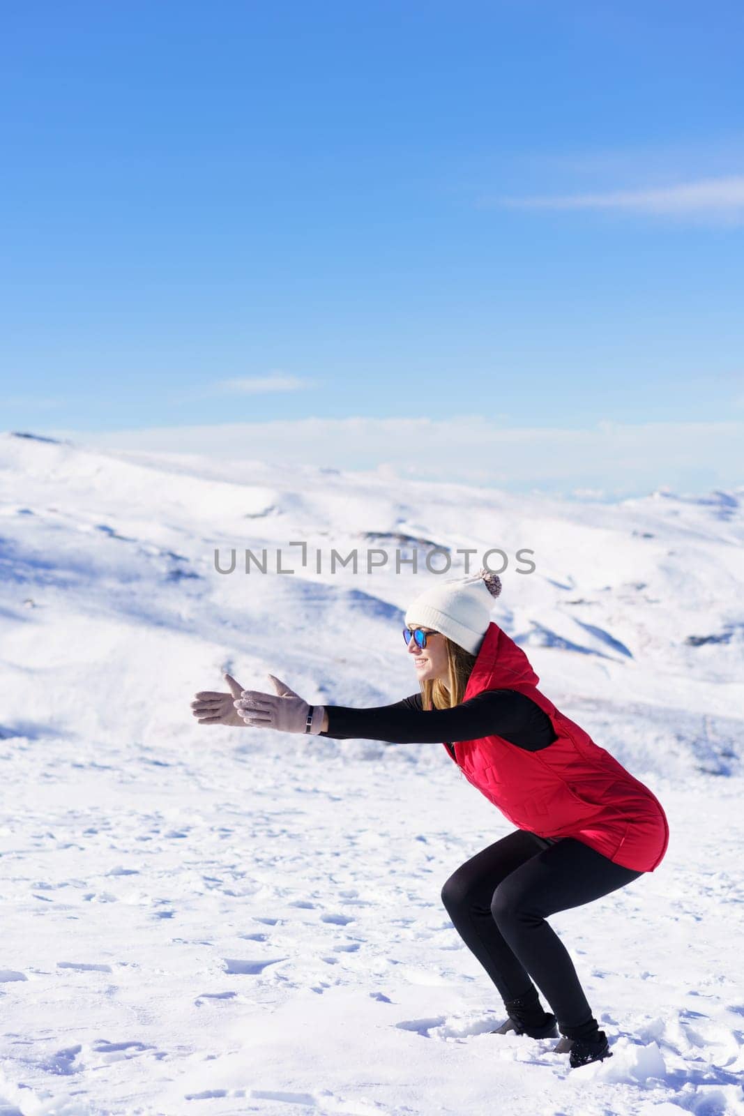 Side view of young female in warm clothes cap sunglasses looking away while squatting with outstretched arms against blue sky and snowy mountains in daylight