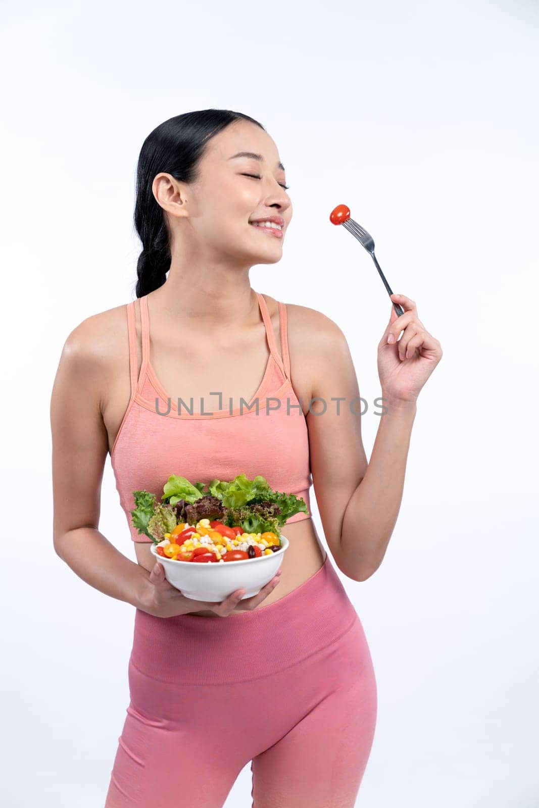 Young sporty Asian woman in sportswear holding salad bowl fill with vibrant of fruit and vegetable. Natural youthful and fit body lifestyle with balance nutrition on isolated background. Vigorous