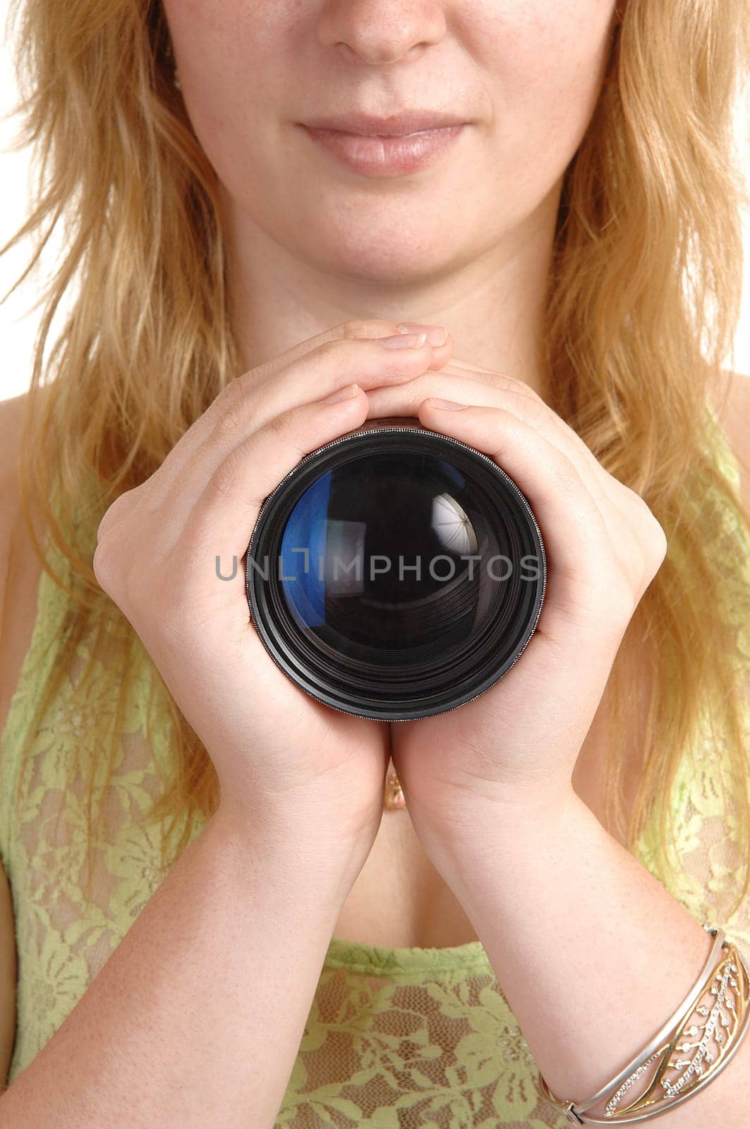 A blond young woman holding a photo lens with both hands for her chest, with reflection in the lens.