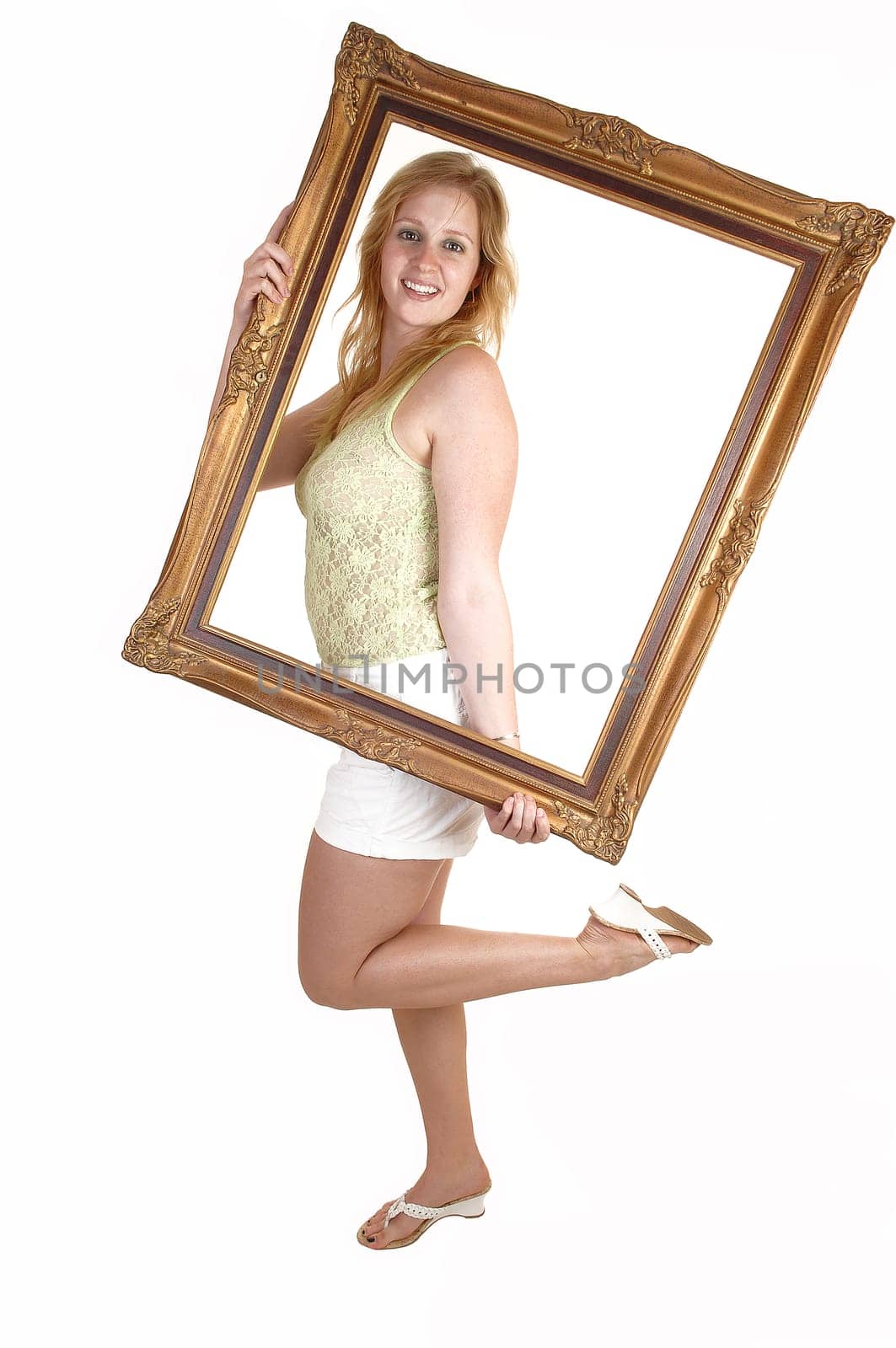 A blond pretty woman in heels and a white shorts and green top, holding a picture frame in front of her, standing for white background.